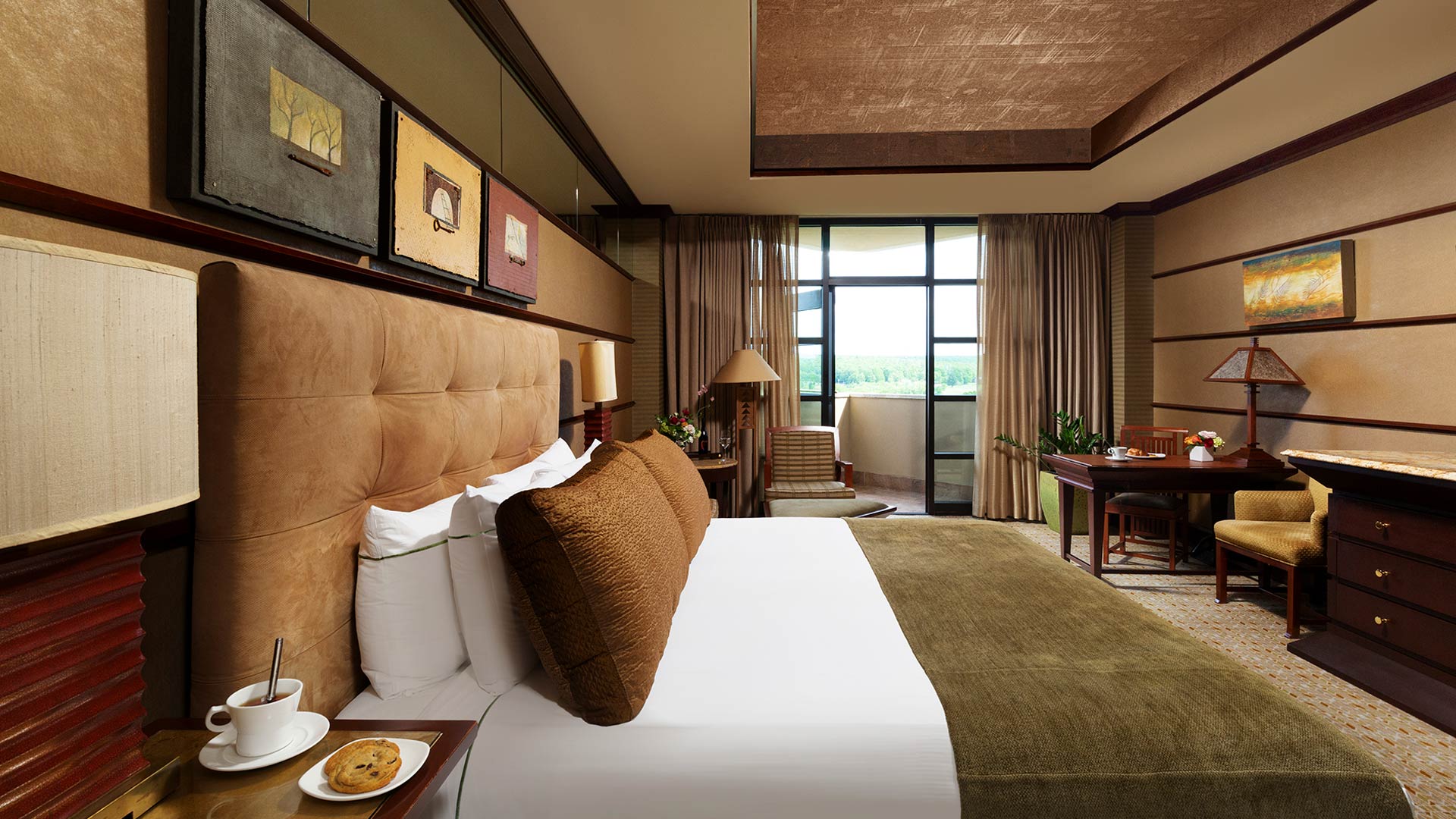 an interior shot of Falling Rock's balcony king room. There is a bed with a plush headboard and white, green and brown bedding. There is a sitting area with a table and dresser across from the bed. There is a door that leads to a balcony overlooking the resort grounds.