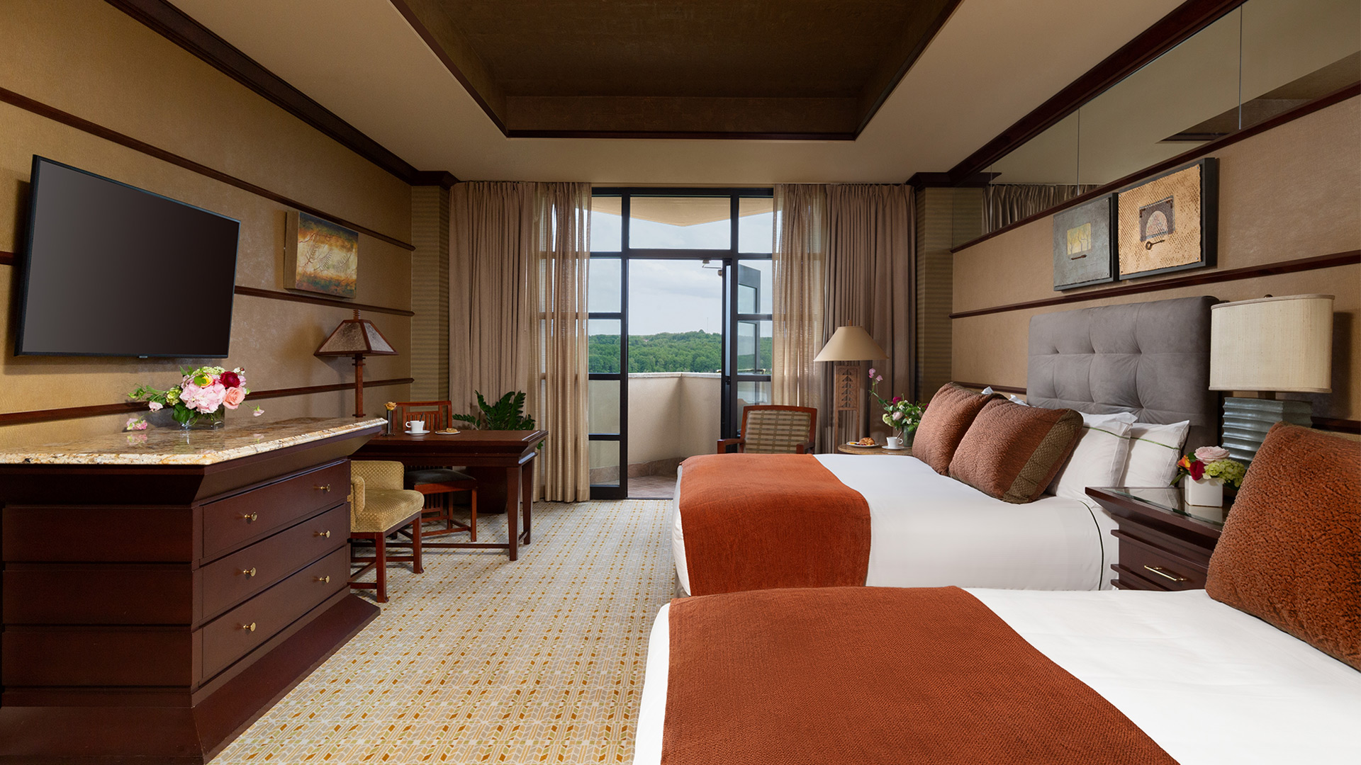 interior shot of Falling Rock's balcony double room. There are two king beds with plush headboards and white, burnt orange and brown bedding. There is a sitting area and a dresser with a flatscreen TV mounted above it. There are doors that lead to a balcony overlooking the resort grounds.