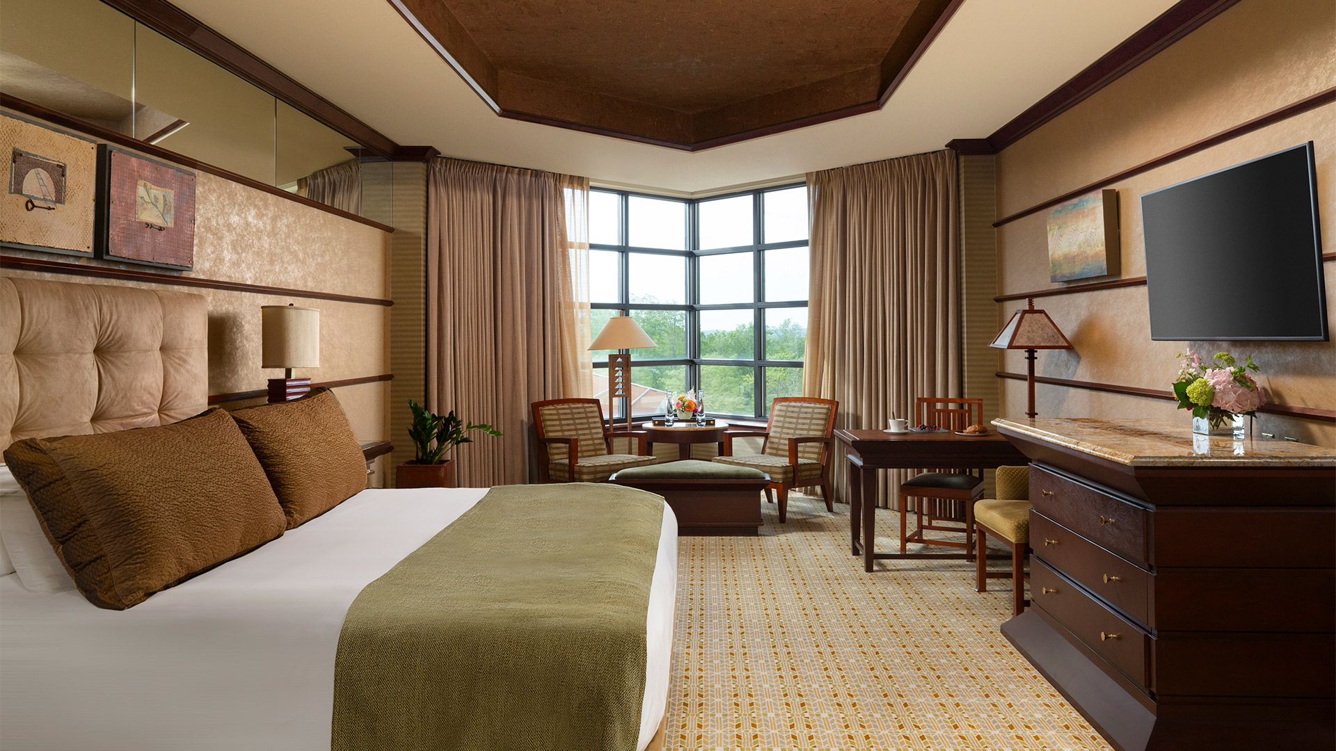 an interior shot of Falling Rock's luxury king room. There is a bed with a plush headboard and white, green and brown bedding. There is a sitting area with a table and dresser across from the bed. There are windows overlooking the resort grounds.