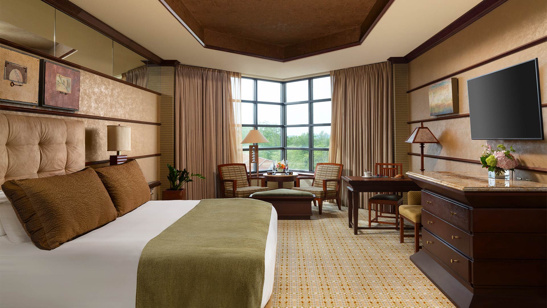 This is an interior shot of Falling Rock's luxury king room. There is a bed with a plush headboard and white, green and brown bedding. There is a sitting area with a table and dresser across from the bed. There are windows overlooking the resort grounds. Next Slide for other carousel items.