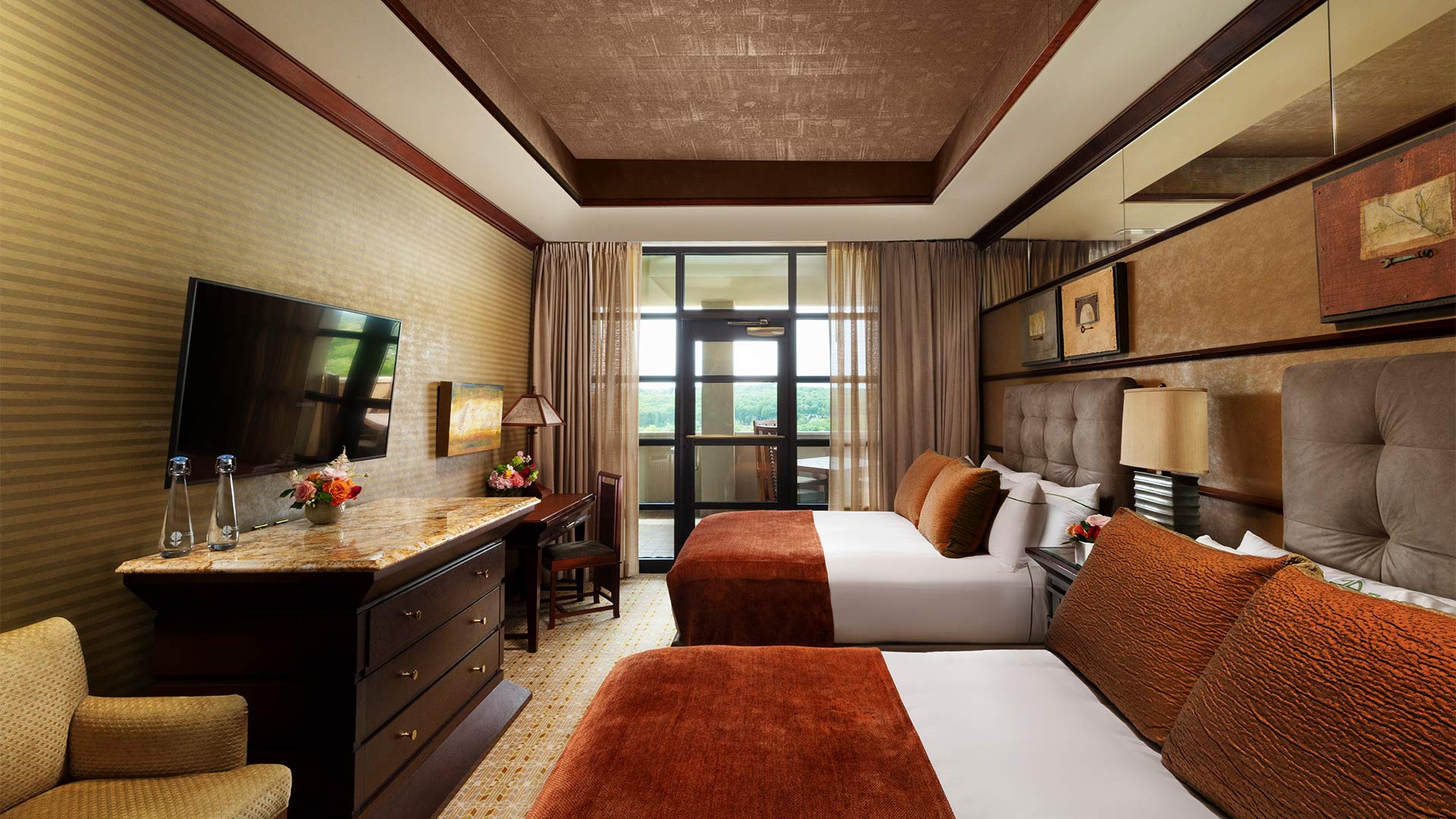 an interior shot of Falling Rock's double suite's bedroom. There are two queen size beds on the right side of the room with white and burnt orange bedding. Across from the beds is a desk and a dresser with a flat screen tv mounted above it. There is a door that leads out to a balcony that overlooks the resort grounds