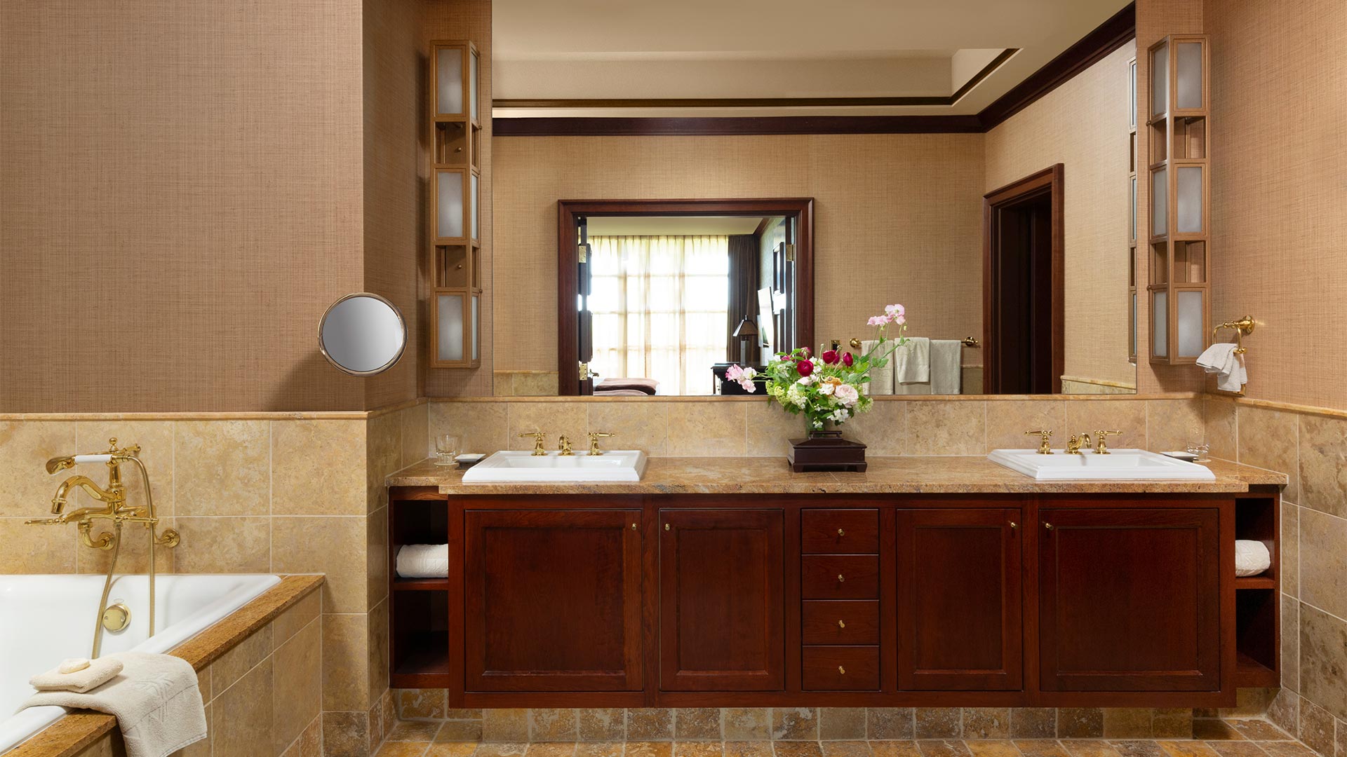 an interior shot of the bathroom. There is a large counter with a sink and shelving. There is a vase full of colorful flowers on the counter top. To the left of the sink is a white whirlpool bathtub with gold hardware.