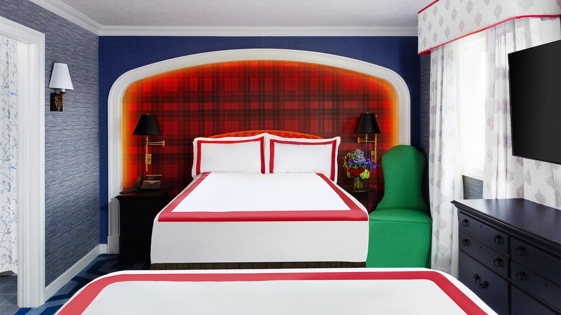 detail shot of a bedroom. The bed has white bedding with bright red piping. There wall behind the bed is a classic buffalo plaid wallpaper. There are blue and green accents throughout the room.