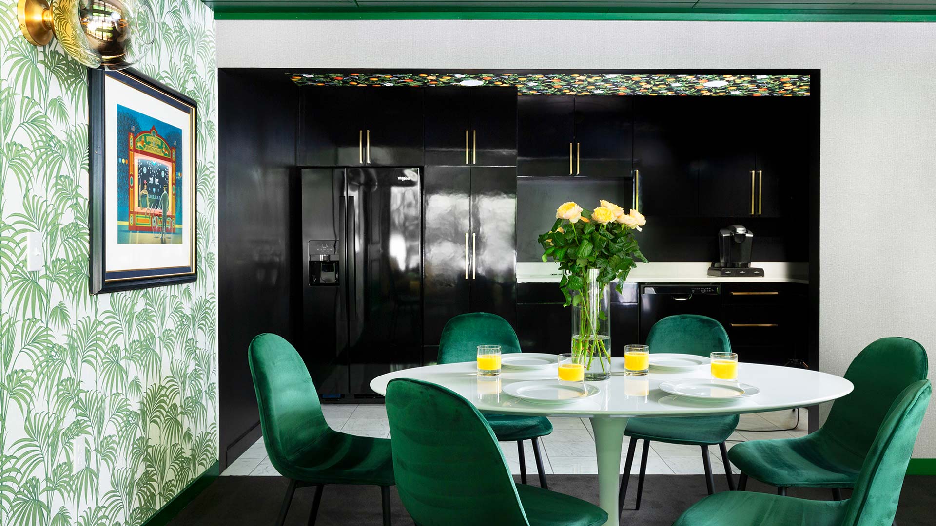 an interior shot of the dining area. There is a large circulat table with glasswear and a floral centerpiece with emerald green chairs surrounding it. In the background is the kitchen.