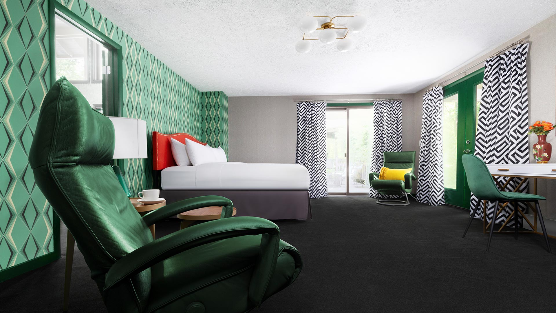 an interior shot of a bedroom. There is an emerald green chair in the foreground. There is a bed with white linens and red headboard. There are sliding doors leading out onto a patio and another green chair next to the doors.