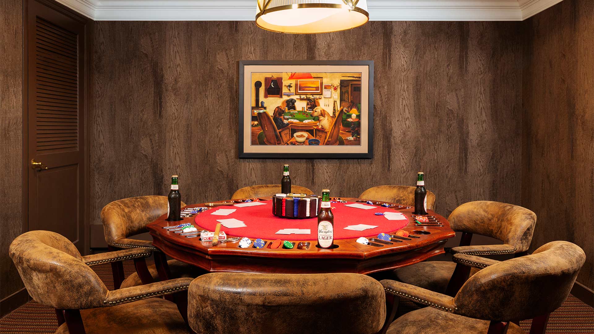 interior shot of a card room. The room has dark wood panels on the walls and the poker table is round and red. There are several plush chairs around the table