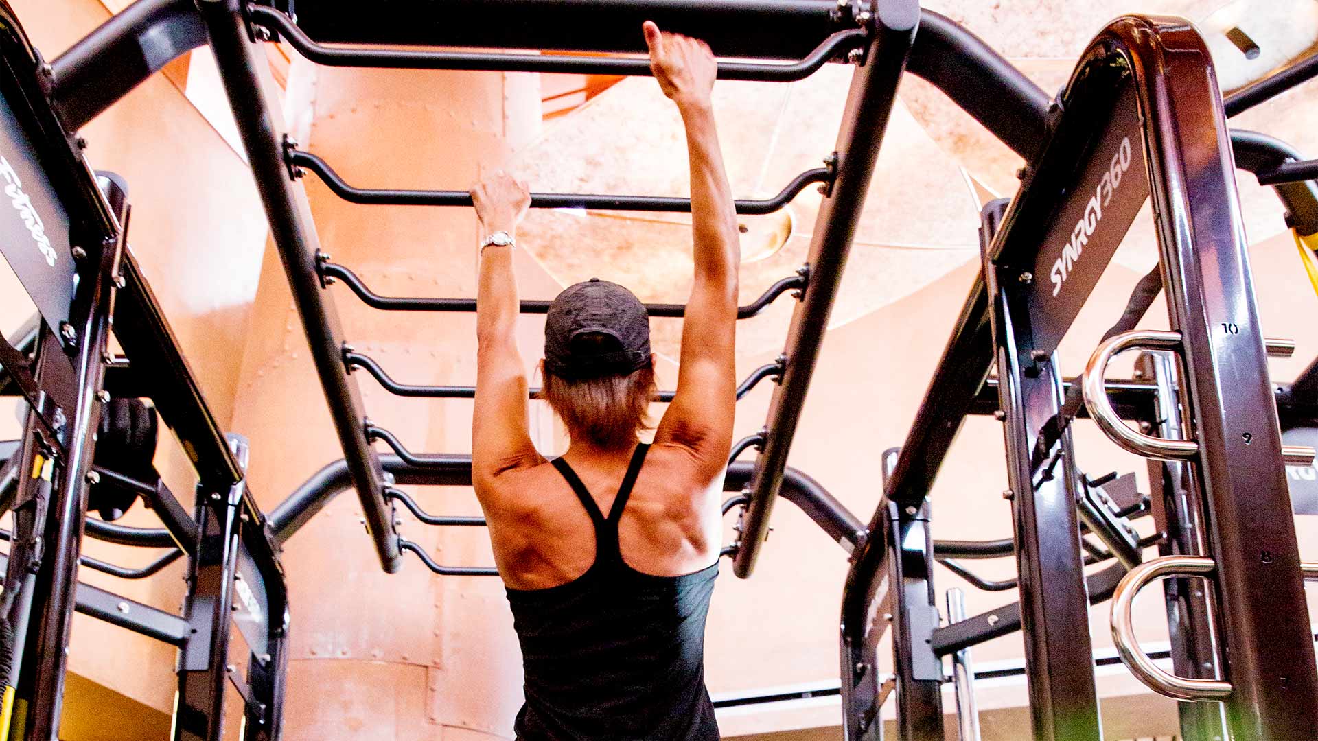 a woman in a black workout tank top and hat is working her way across rungs like monkey bars