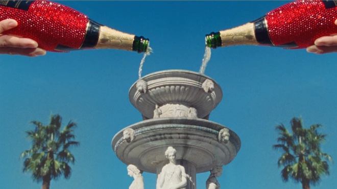 two red jewel bedazzled champagne bottles are being poured into a greek-inspired two-story fountain. There is a blue sky and palm trees in the background