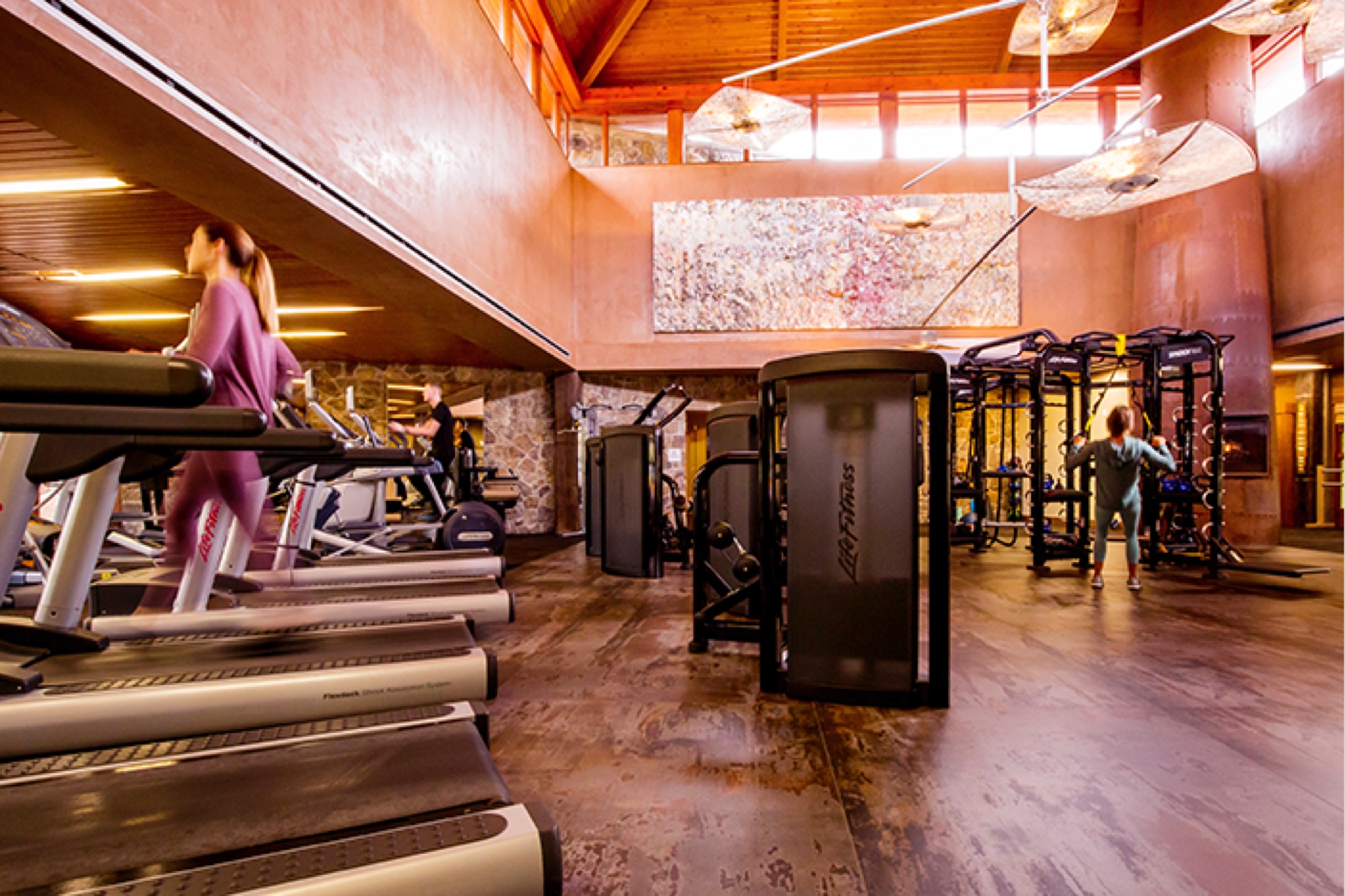 an action shot of a fitness center. There are treadmills lining the left side of the gym. There is a woman walking on one of the treadmills. There are weight machines on the right side of the room with a man using them