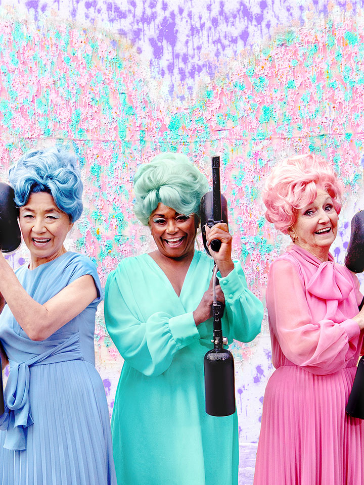 Three older women wearing monochrome outfits with wigs, one blue, one green, one pink, in front of a colorful backdrop