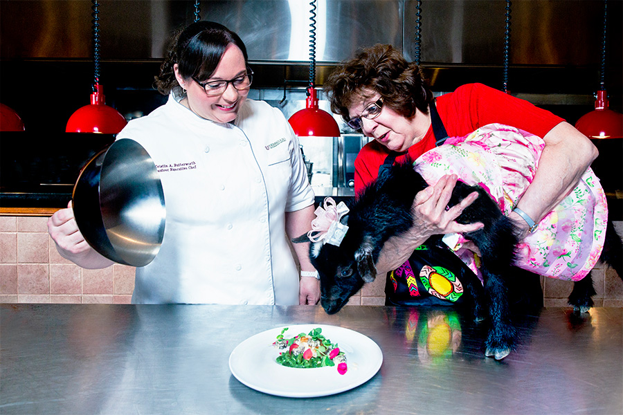 two women, one chef unvailing a plate of food and the other holding a black goat in a dress up to the plate