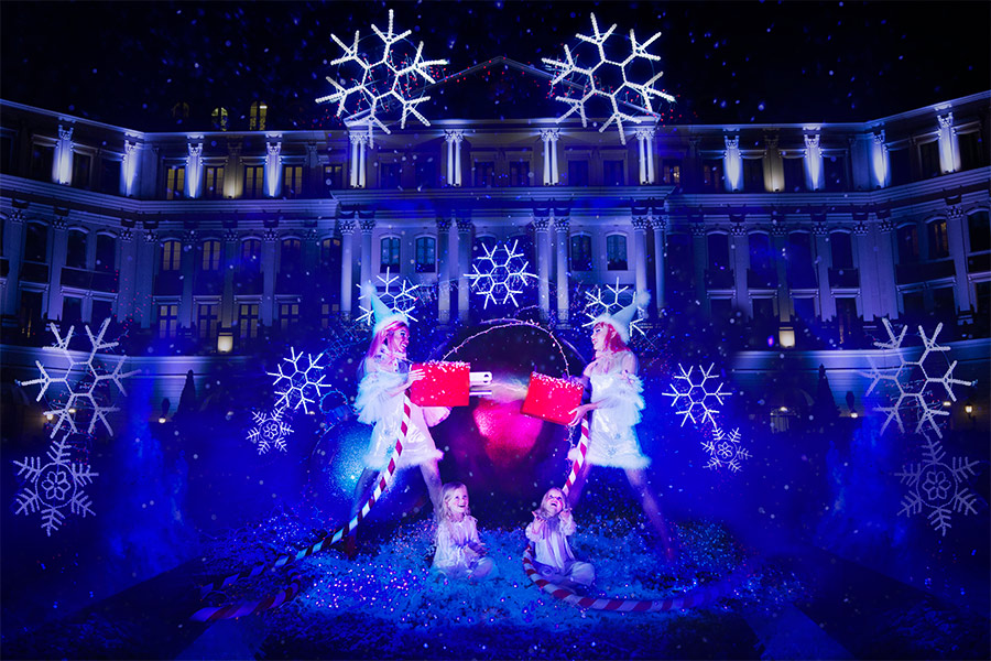 Christmas light setup with glowing snowflakes in front o fa large building and two people dressed as elves in the front putting together a giant electrical outlet