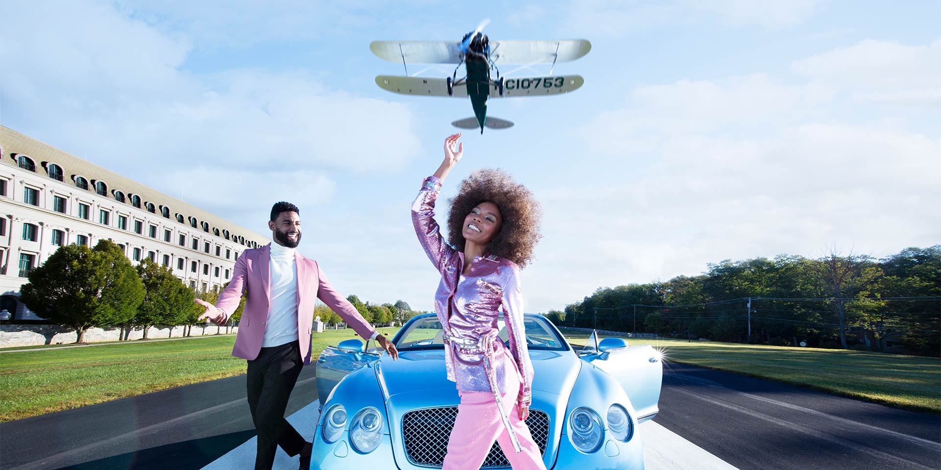 A couple dressed in pink attire walking near a blue car with a airplane flying above them.