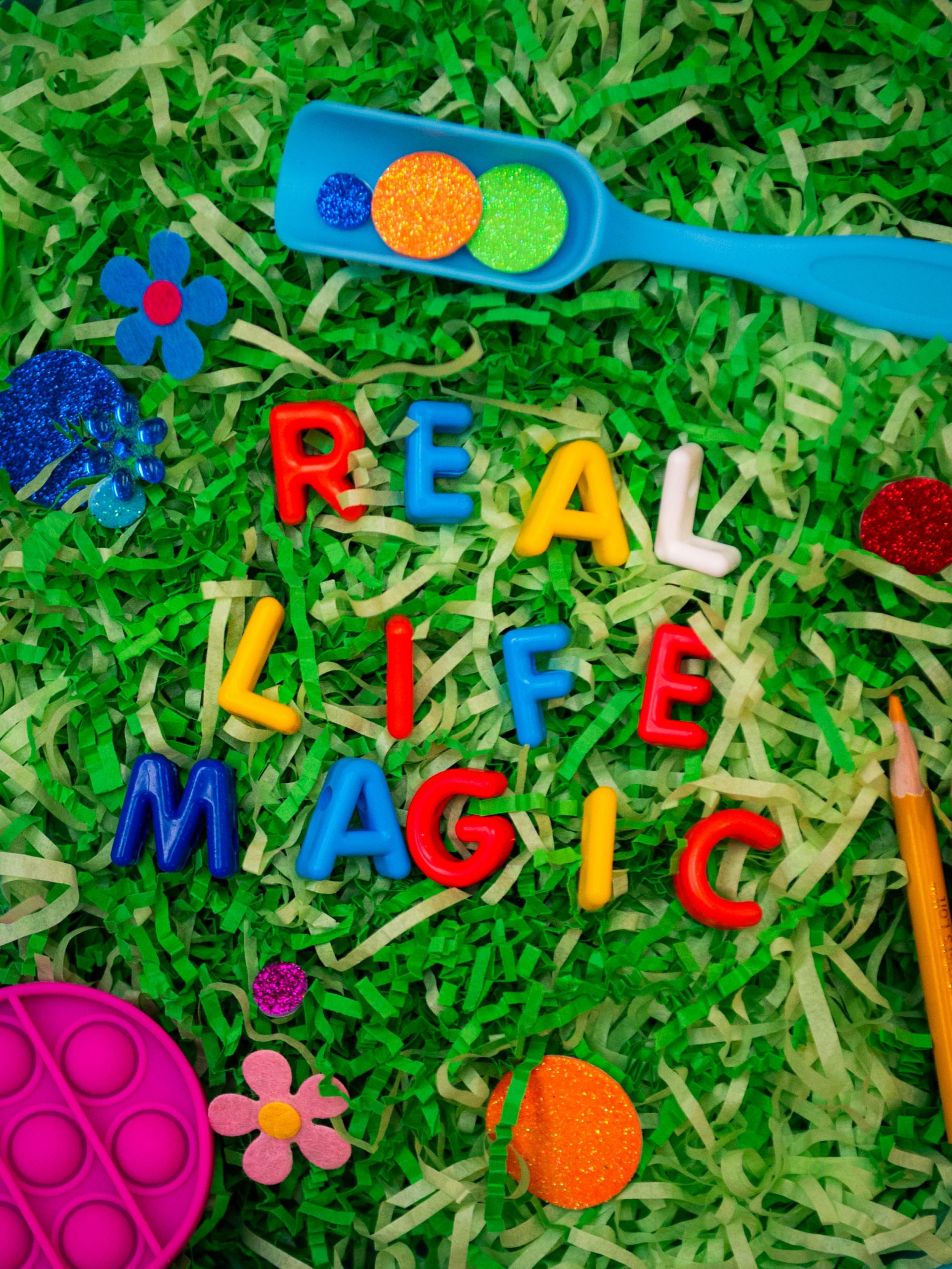 The words "Real Life Magic" are spelled out with colorful letters placed on top of fake grass.