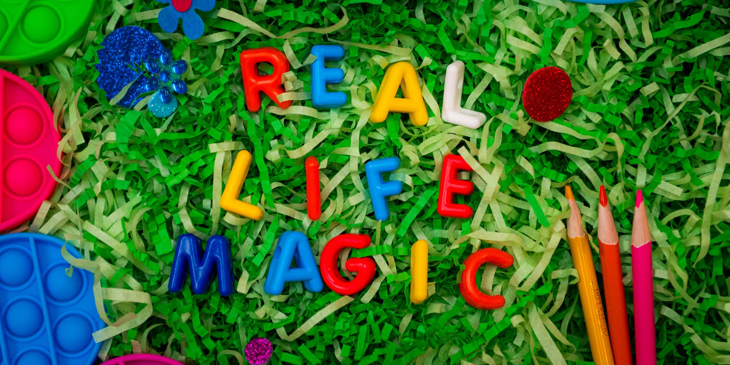 The words "Real Life Magic" are spelled out with colorful letters placed on top of fake grass.