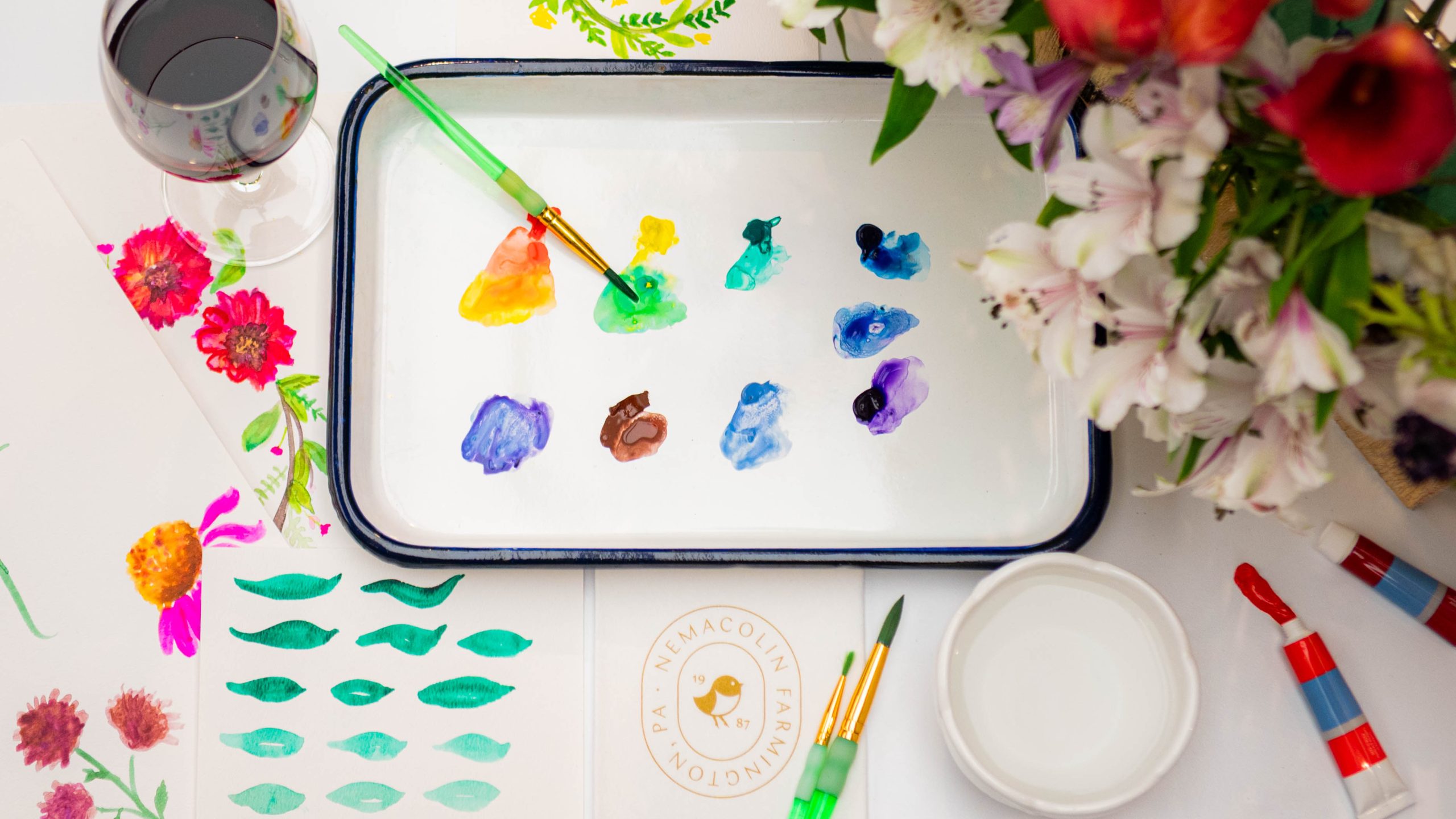 Large wine glass next to tray of watercolor paints with a bird napkin