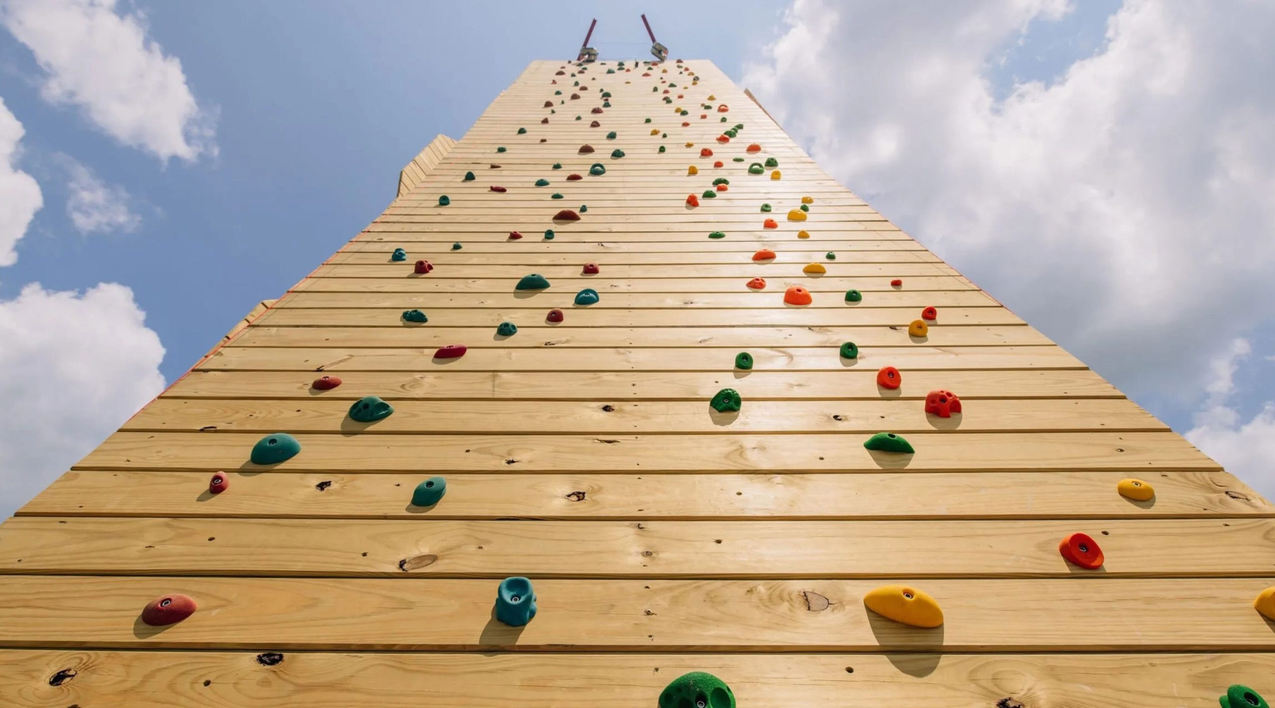 Wooden climbing wall reaching into a blue sky with white clouds showing colorful foot holds and handholds