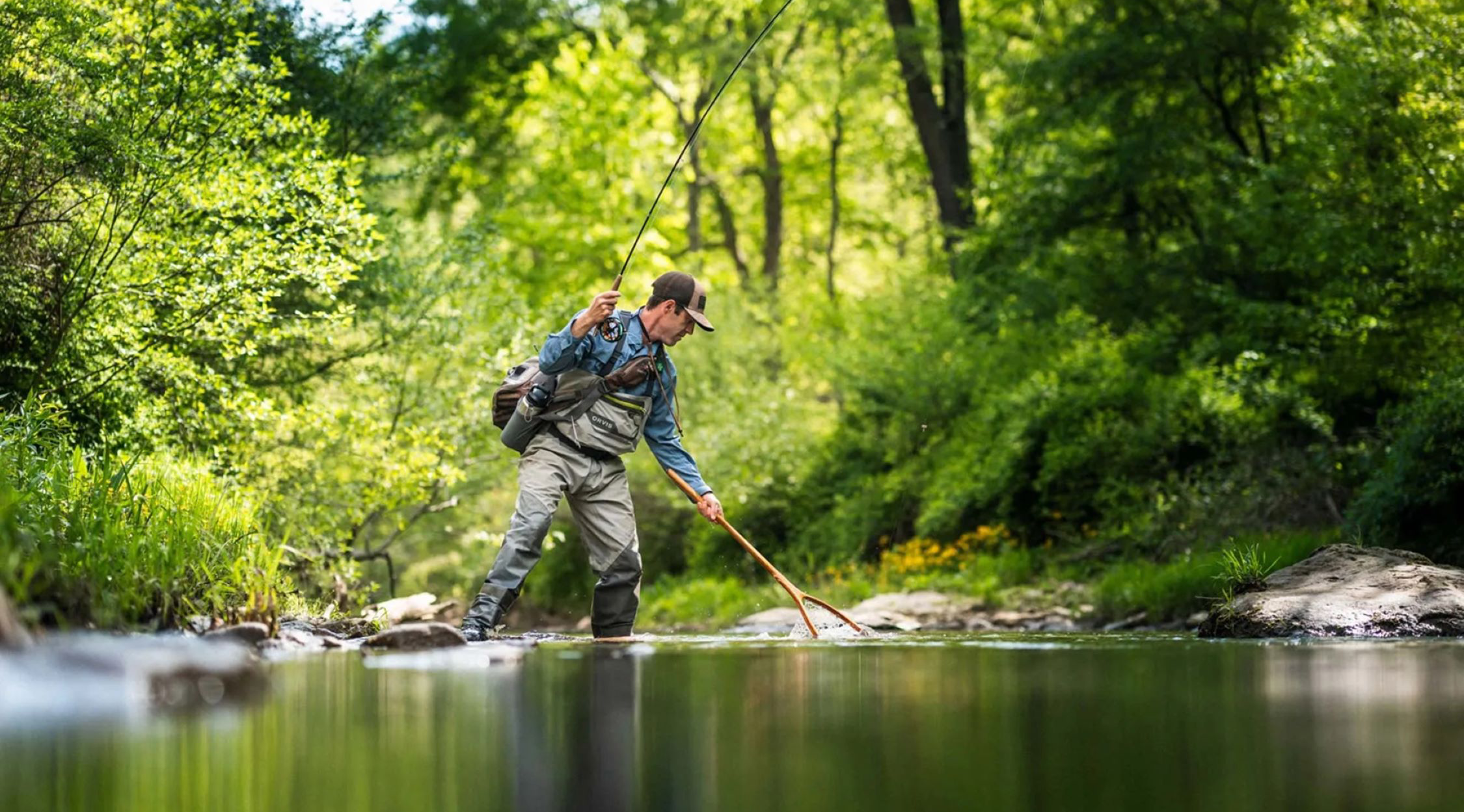 A male fly fishing with a spin rod and net, wearing waders, and in a wooded stream