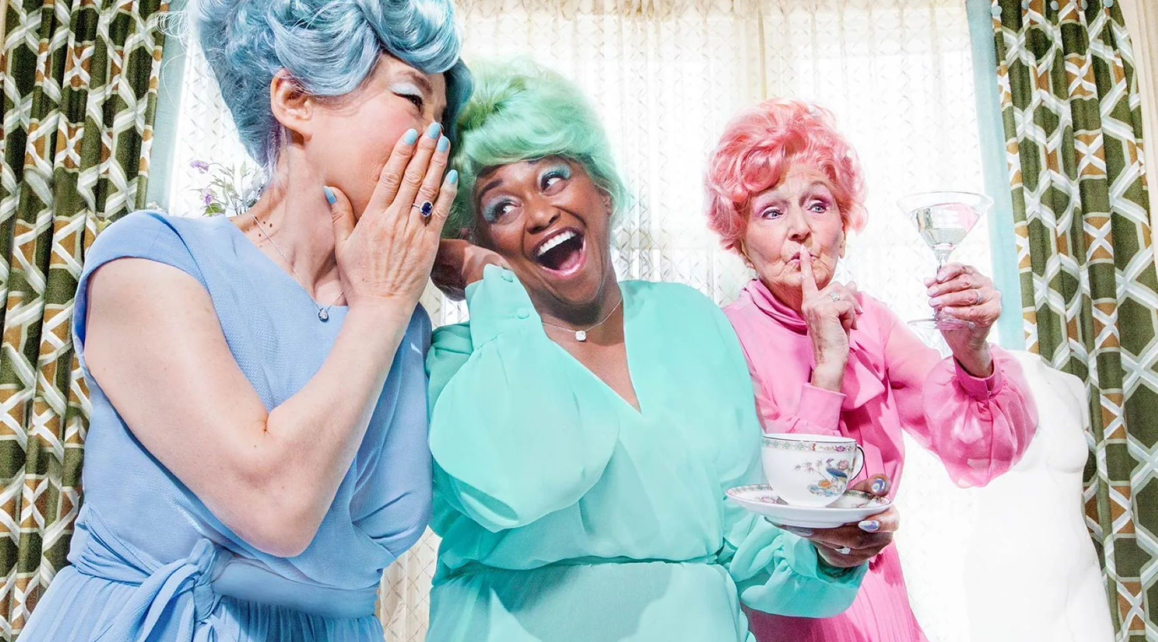 Three women in colorful pastel dresses with matching wigs of blue, green, and pink, laughing and sharing a secret.