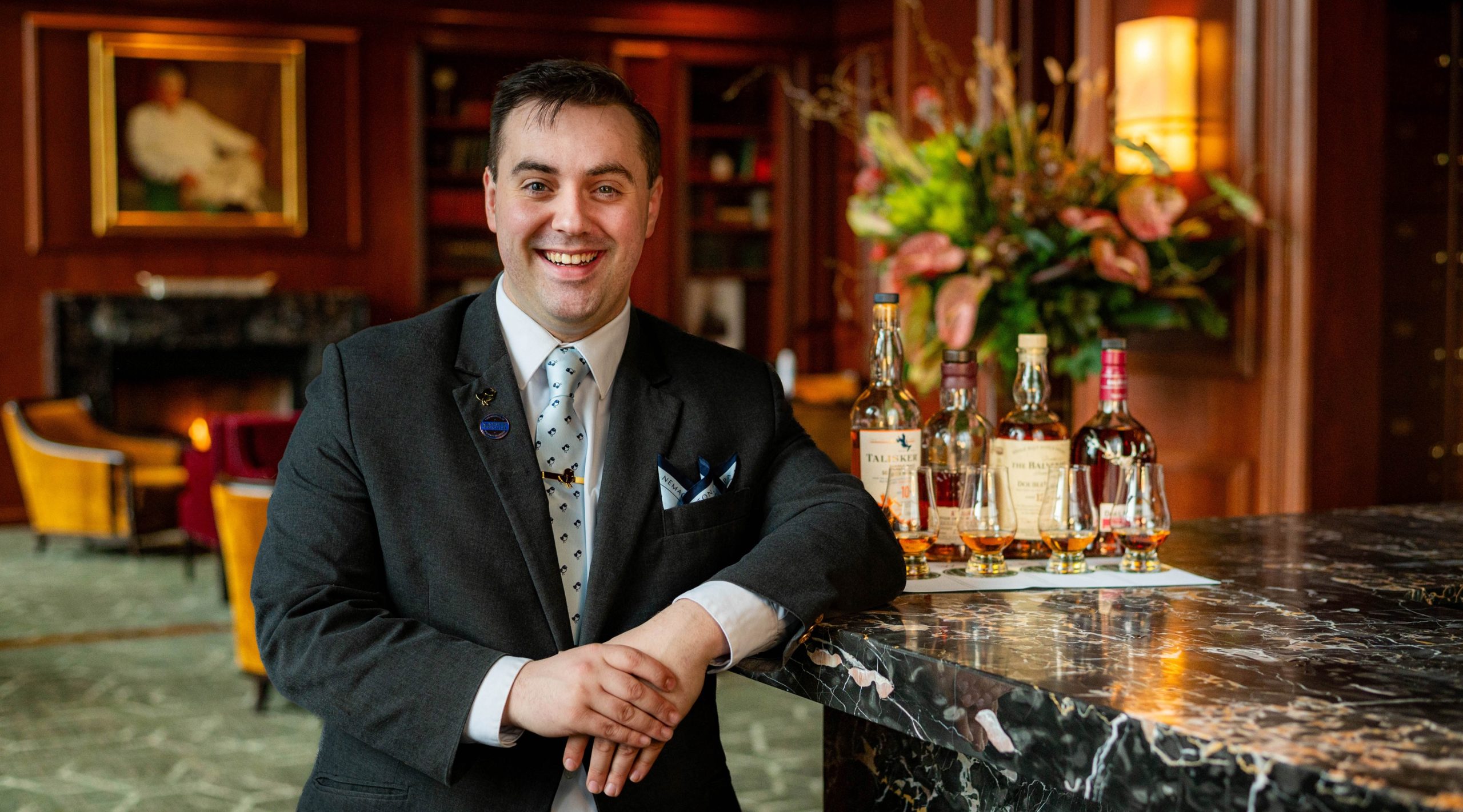 A man wearing a dark suit and a light blue tie smiles while leaning against a bar top. Next to him are four whiskey bottles and four glasses.