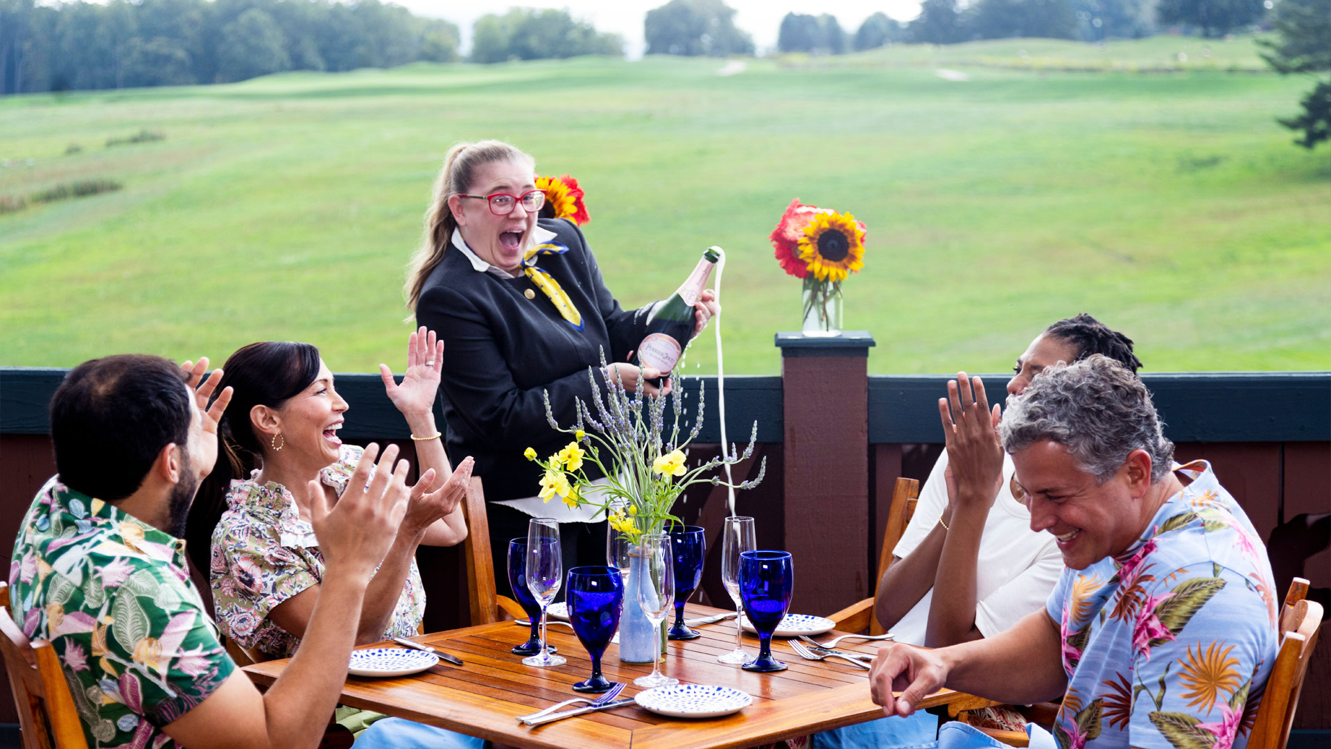 A woman popping a bottle of champagne at a table of four people who are seated at outdoor seating on a deck overlooking a green landscape
