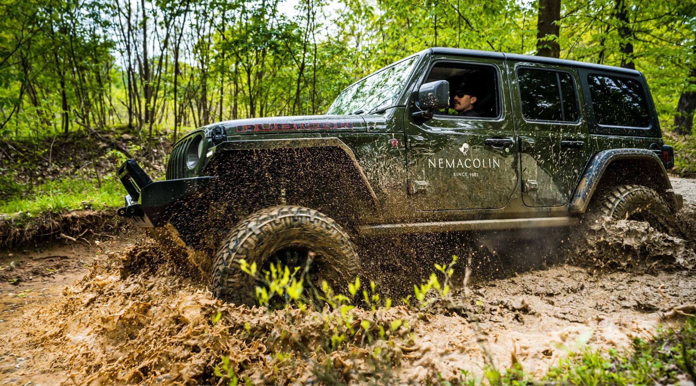 A green Jeep with a white Nemacolin logo on the door drives through mud in the woods.