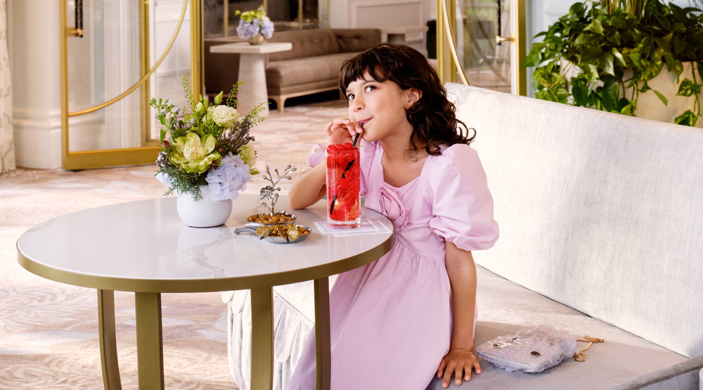 A girl wearing a pink dress drinks a Shirley Temple beverage through a black straw. She is looking up while sitting in the Lobby Bar at Nemacolin.