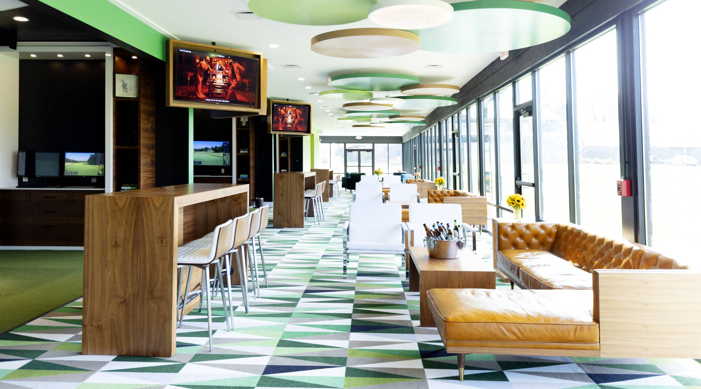 A colorful scene with green carpet, brown sofas and tables, and televisions next to bays of golf simulators at The Golf Academy at Nemacolin.